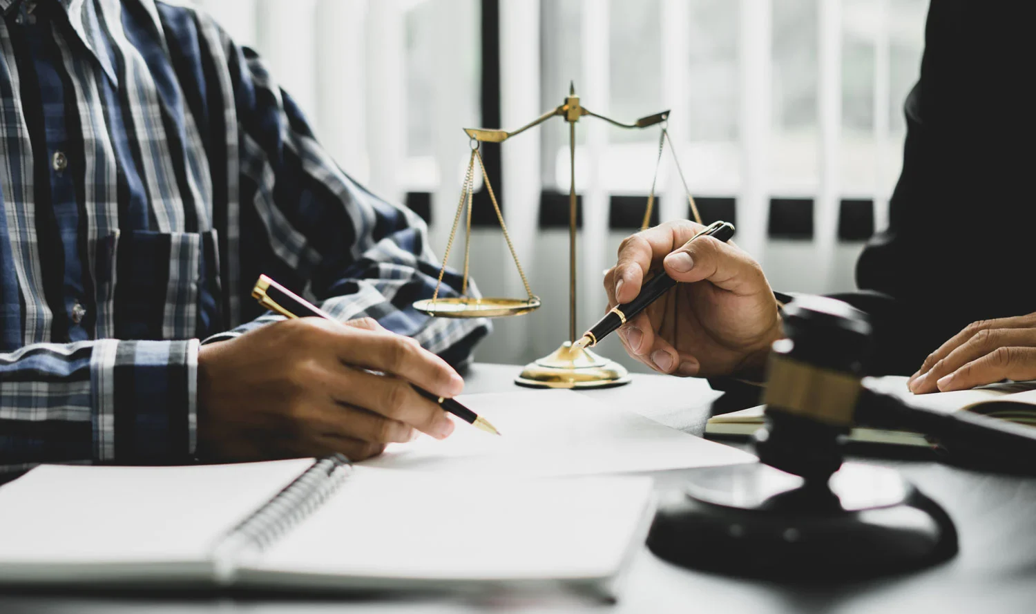 Two individuals meeting at a table, one in a plaid shirt holding a pen, with a gavel and scales of justice in view